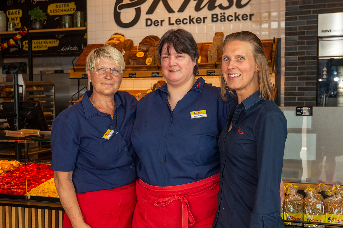 Karina Moritz, Geschäftsführerin Hanna Kruse und eine weitere Mitarbeiterin von Bäcker Kruse stehen vor der Bäckerei-Theke in Arbeitskleidung und lächeln