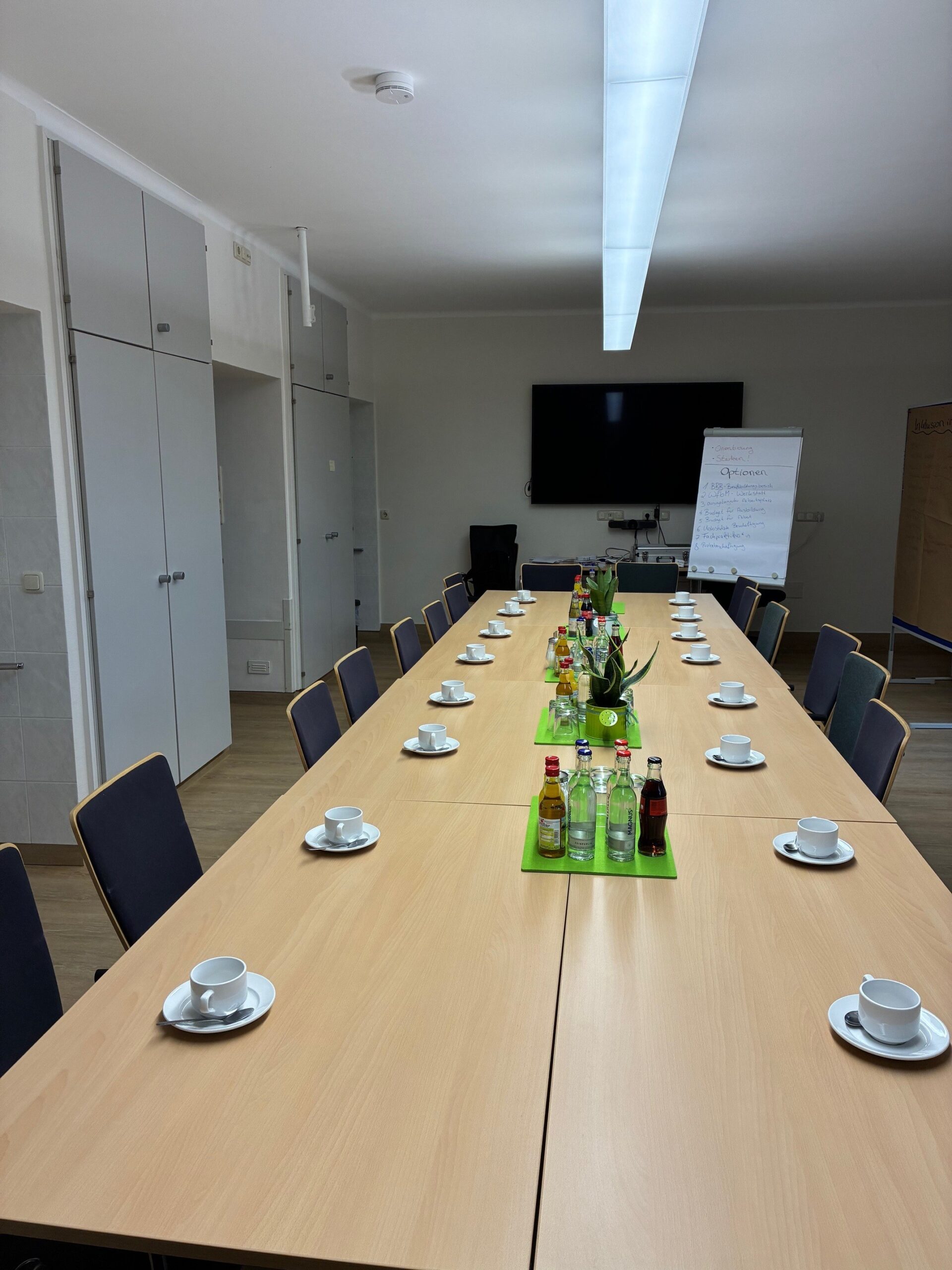 Foto vom Workshop Entgeltsystem in Werkstätten für behinderte Menschen, ein gedeckter Tisch für 16 Teilnehmende und ein beschrifteter Flipchart in einem Besprechungsraum